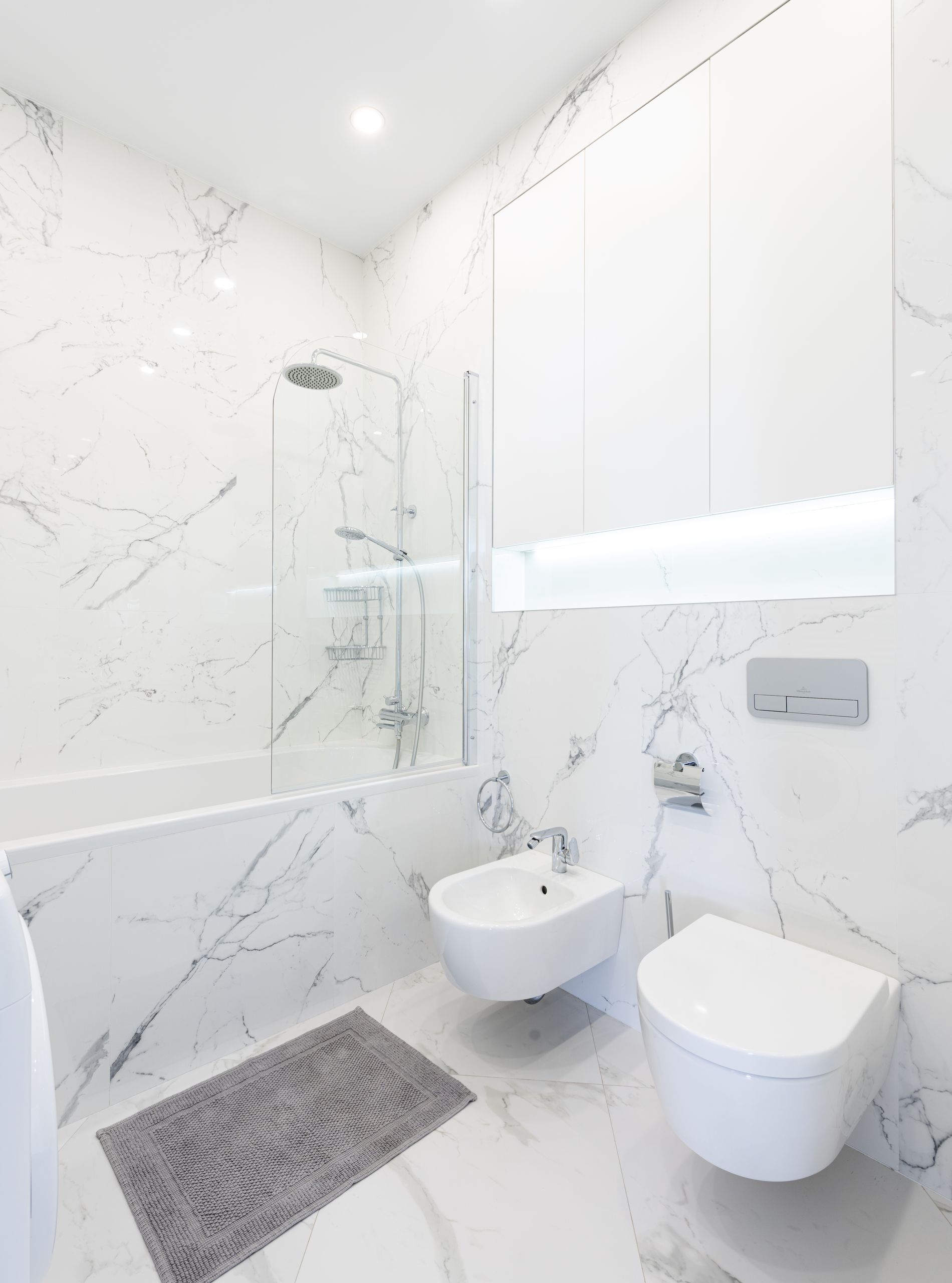 Interior of light bathroom with toilet and bidet under mirror on white wall next to bathtub with shower near carpet on floor